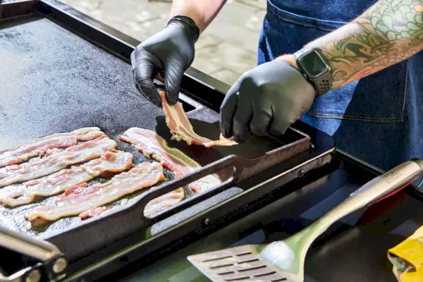 Una persona coloca tocino en la parrilla Traeger Flatrock Flat Top Grill