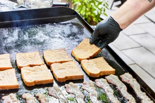 Una persona cocina tostadas francesas y tocino en la parrilla de gas Blackstone 1554 Cooking de 4 quemadores con parte superior plana