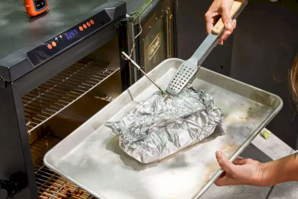אדם מסיר בשר מה-Dyna-Glo 30-Inch Digital Bluetooth Electric Smoker, כסף (DGU732SDE-D)