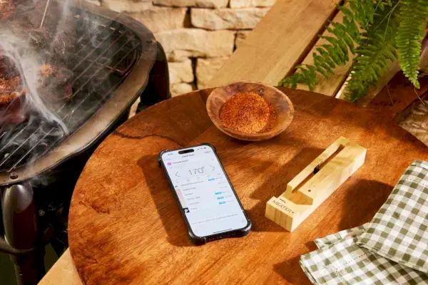 טלפון עם אפליקציית The Meater Plus Smart Meat Thermometer מוצגת על שולחן ליד קערת תבלינים והמטען