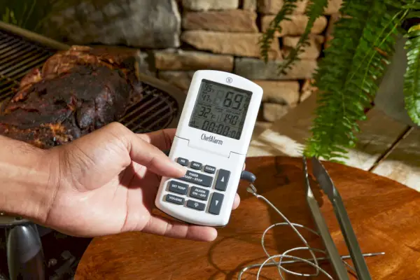 Dłoń trzymająca ThermoWorks ChefAlarm obok drewnianego stołu i grilla
