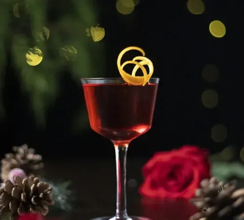 červený kokteil v kokteilovom pohári nick a nora s oranžovým zákrutom' title=