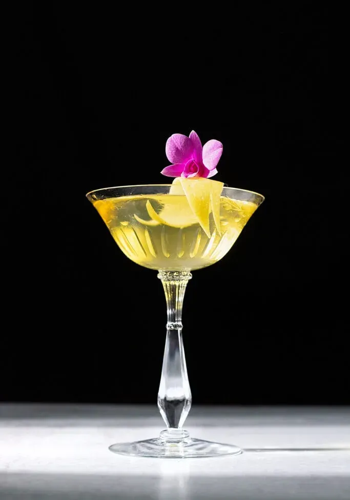 Leuchtend gelber Cocktail in einem Coupé-Glas mit rosa Blume.' title=