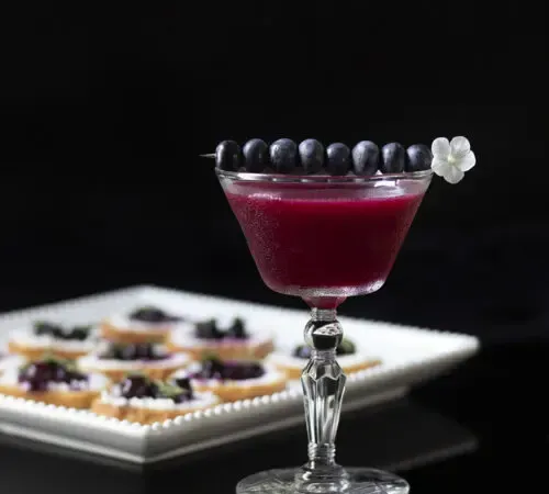 dunkelvioletter Cocktail neben einer Platte mit Vorspeisen' title=