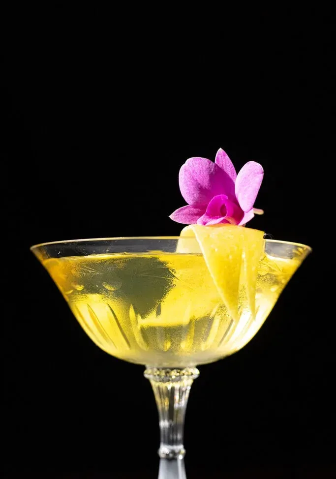 cóctel amarillo con cáscara de limón y orquídea rosa.' title=