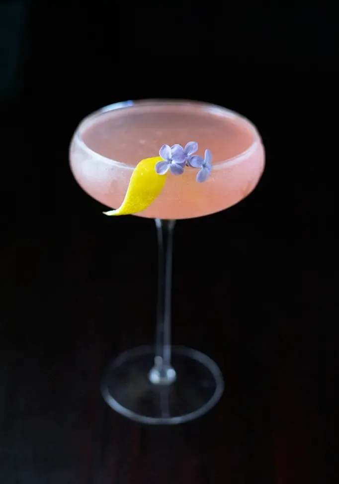 vista de arriba hacia abajo de cóctel rosa con enfoque superficial en la guarnición de flores de lila' loading='lazy' title=