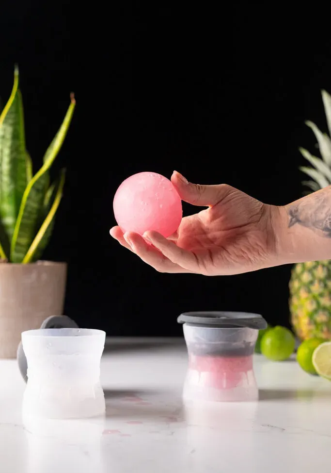 tenendo una grande sfera di ghiaccio rosa accanto agli stampini per il ghiaccio.' loading='lazy' title=