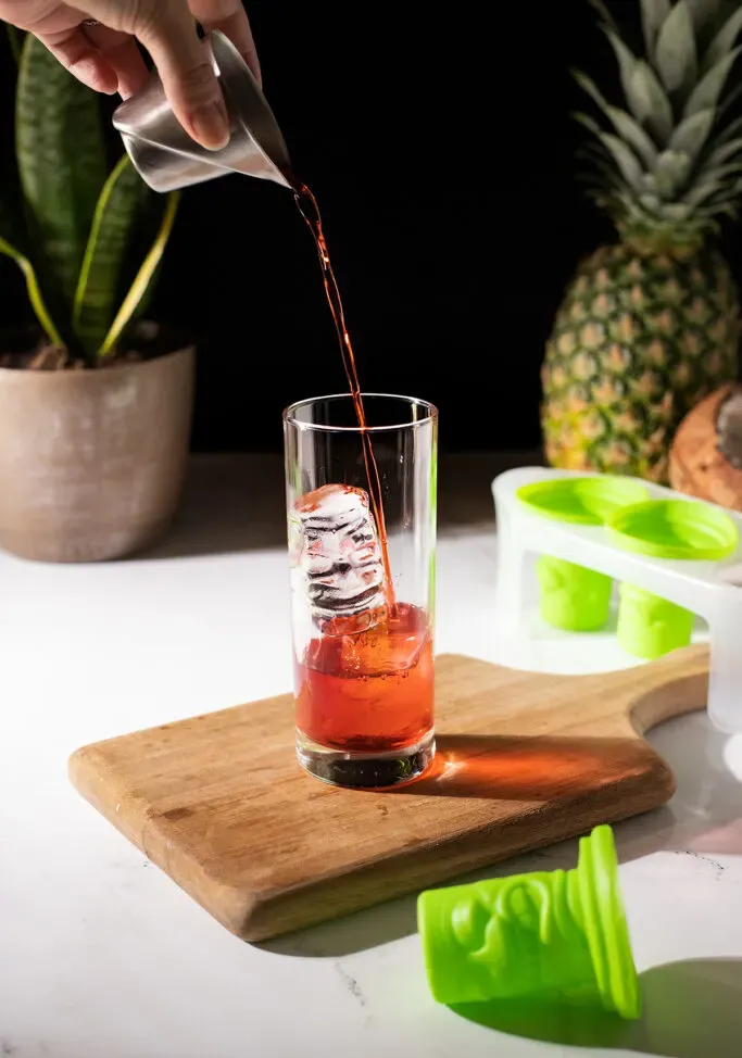 versare il liquore rosso sul ghiaccio tiki trasparente in un bicchiere highball.' title=