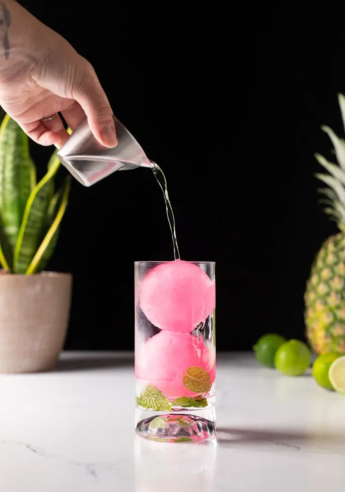 versando il rum bianco in un bicchiere colmo di sfere di ghiaccio rosa.' title=