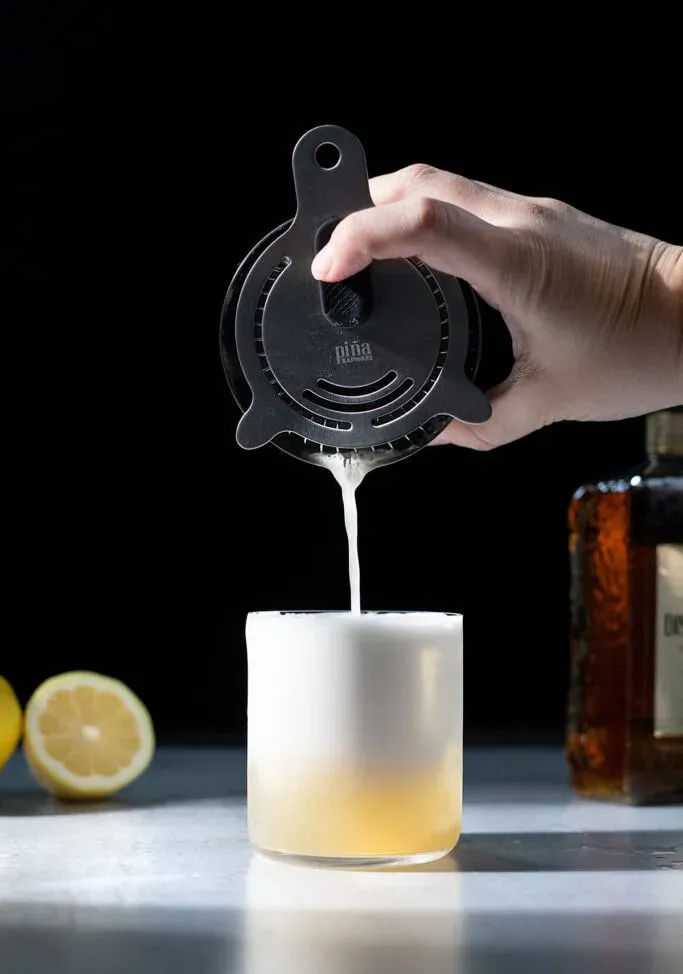 vertiendo un cóctel espumoso en un vaso bajo lleno de hielo.' title=