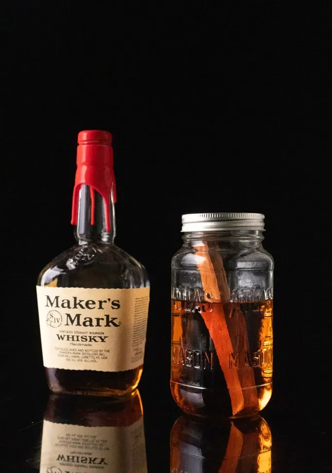 eine Flasche Maker’s Mark Bourbon neben einem mit Bourbon gefüllten Glas und einem Stück Kirschholz.' title=