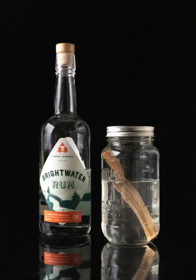 eine Flasche Brightwater White Rum neben einem Glas mit weißem Rum und einem Stab aus Ahornholz.' title=