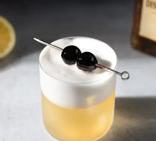 un cocktail doré avec une épaisse mousse blanche et deux cerises.' title=