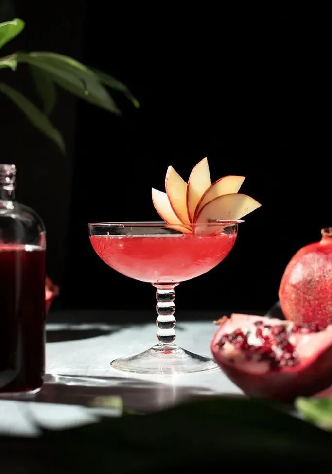 Ein roter Cocktail in einem großen Vintage-Coupé-Glas, garniert mit einem Apfelfächer' title=