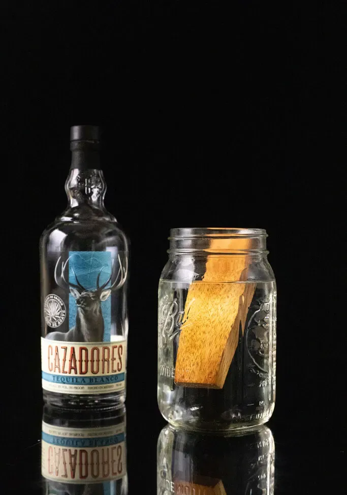 láhev tequily Cazadores blanco vedle zednické nádoby naplněné tequilou a dřevem amburana.' title=