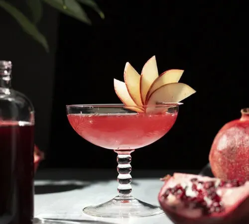 un cóctel rojo en una gran copa vintage adornada con un abanico de manzana' title=