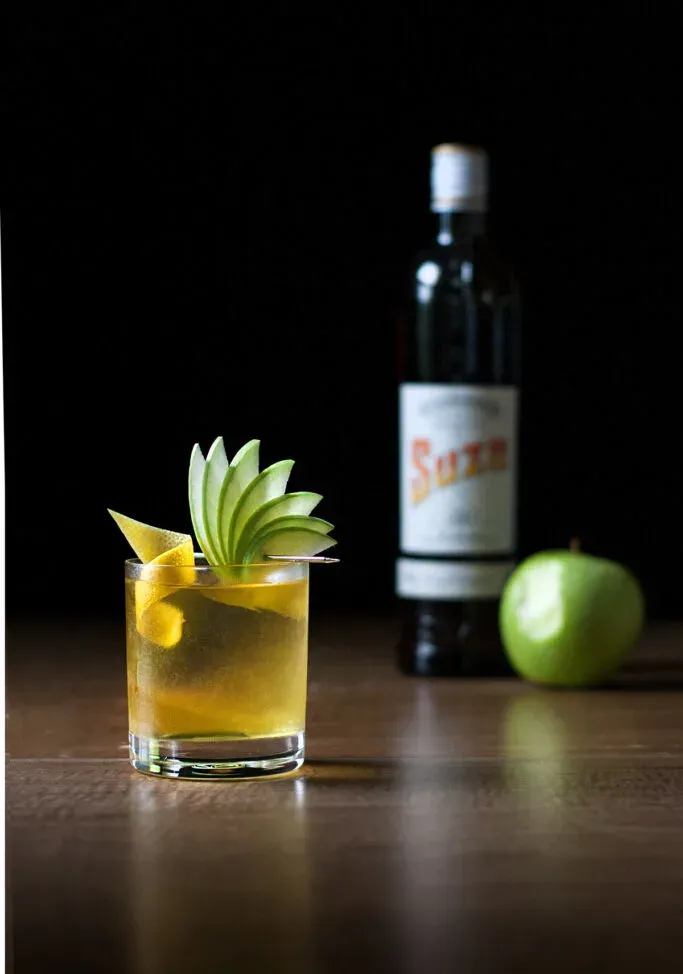 un cóctel amarillo con un abanico de manzana junto a una botella de Suze.' loading='lazy' title=