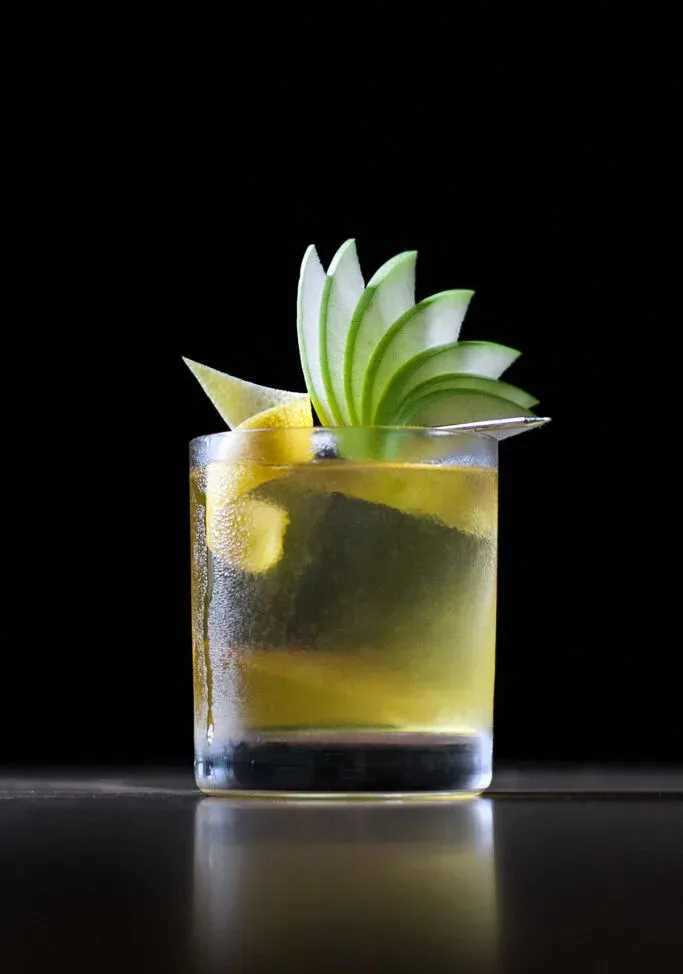 una bebida amarilla en un vaso bajo con un cubito de hielo transparente y una elegante guarnición de abanico de manzana.' loading='lazy' title=