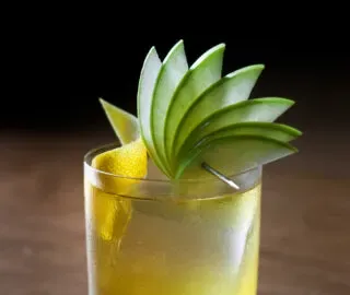 una bebida amarilla en un vaso bajo adornada con una rodaja de limón y un abanico de rodajas de manzana verde.' title=