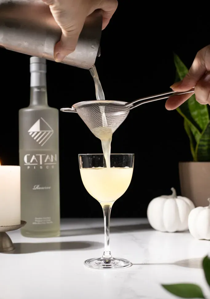 verser un cocktail jaune pâle à travers une passoire à mailles fines dans un verre à cocktail.' loading='lazy' title=