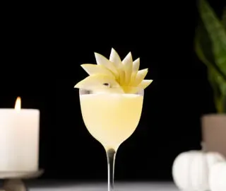 un cocktail jaune pâle dans un verre Nick et Nora avec une garniture en éventail de poire.' title=