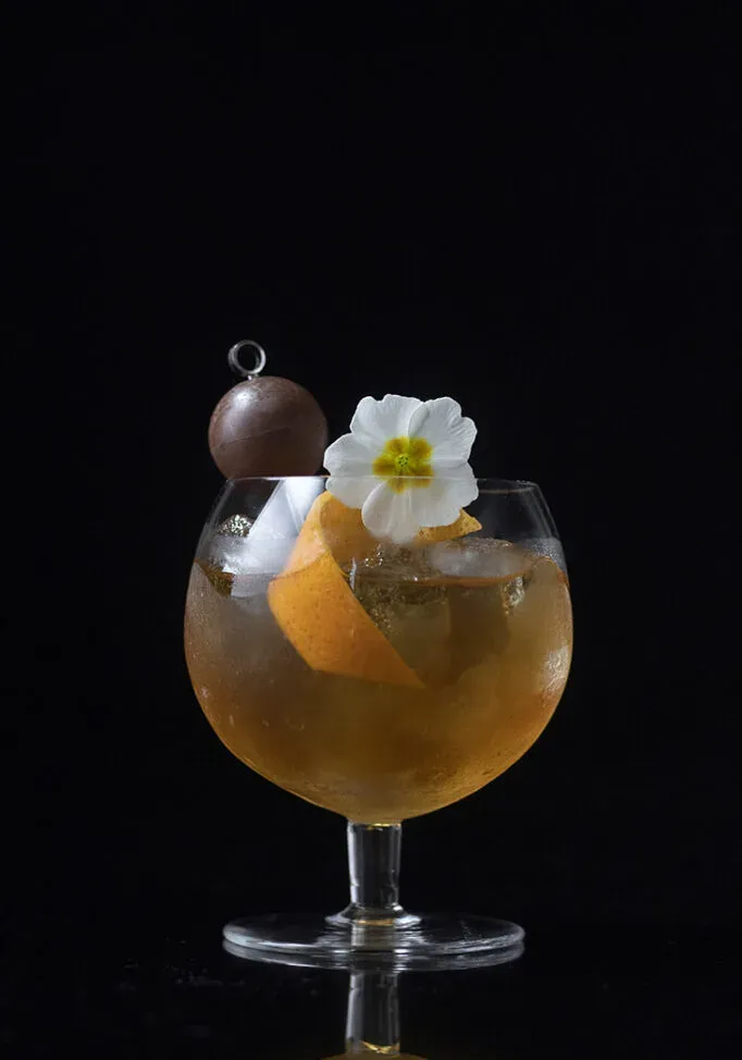 un cocktail au cognac dans un petit snifter avec un zeste d'orange à la truffe au chocolat et une fleur blanche.' loading='lazy' title=