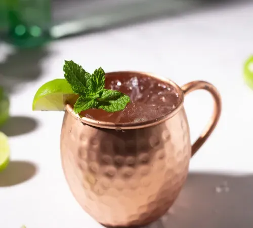 Kupfer-Mule-Becher, gefüllt mit eisiger Flüssigkeit und garniert mit Minzzweig und Limettenspalte.' title=
