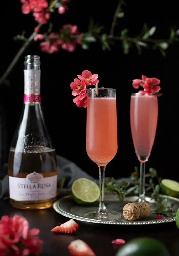 deux cocktails roses finis dans des flûtes à champagne garnies de fleurs de coing' loading='lazy' title=