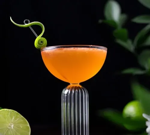pomarańczowy koktajl w kieliszku coupe z fantazyjnym dodatkiem skórki limonki.' title=