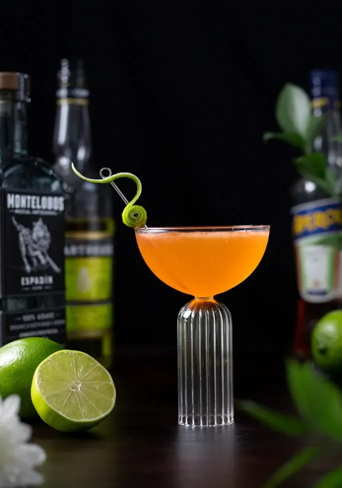 pomarańczowy koktajl w szklance coupe obok butelek z alkoholem.' title=