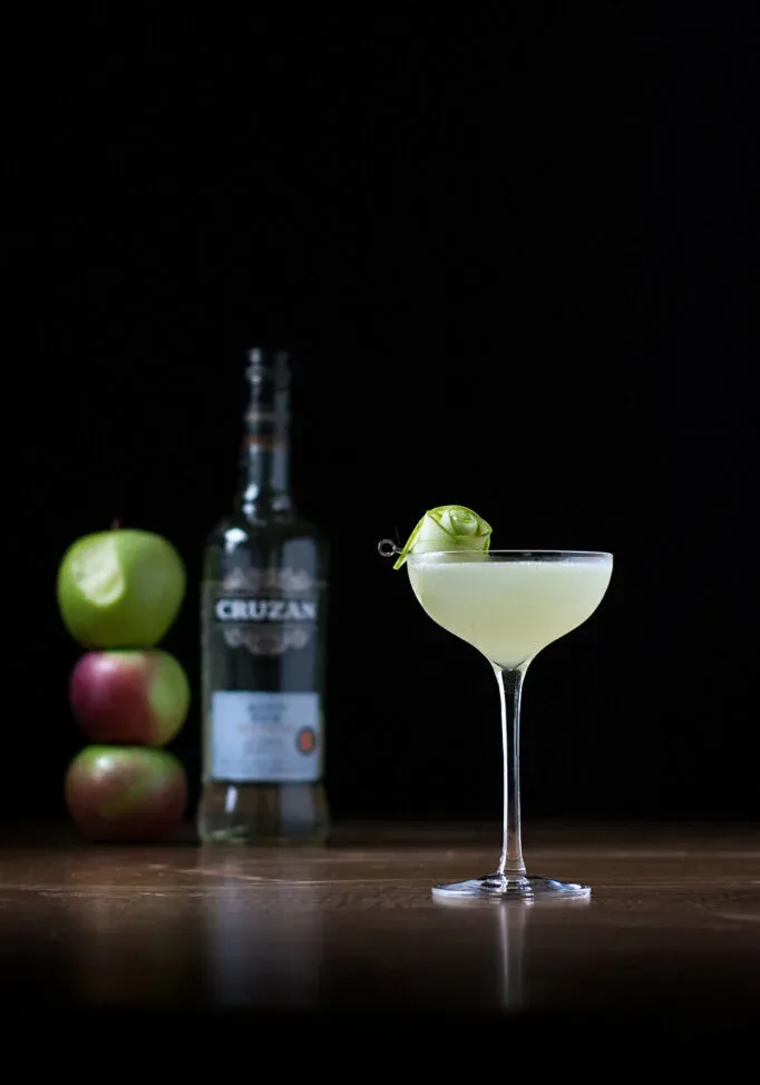 un cocktail in un bicchiere da coupé accanto a una bottiglia di rum bianco e tre mele impilate.' loading='lazy' title=