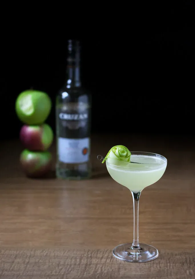 un cocktail verde pallido accanto a una bottiglia di rum bianco Cruzan e tre mele.' loading='lazy' title=