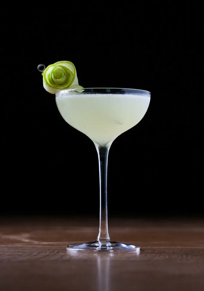 ein blassgrüner Cocktail in einem Coupéglas, garniert mit einer Rose aus grünen Apfelscheiben.' title=