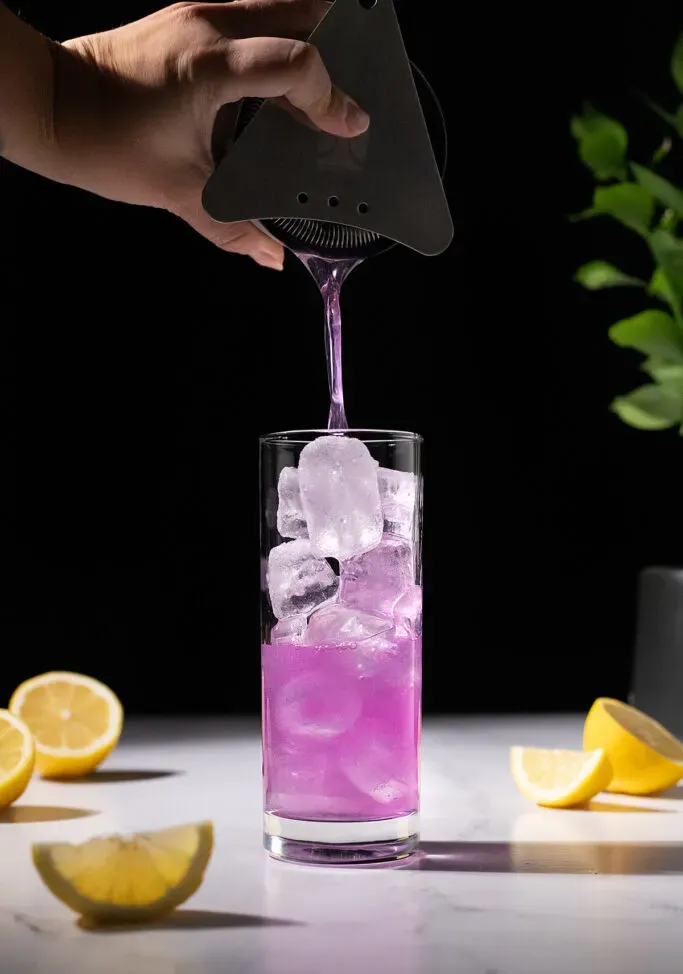 Einen violetten Cocktail in ein hohes, mit Eis gefülltes Glas abseihen.' title=