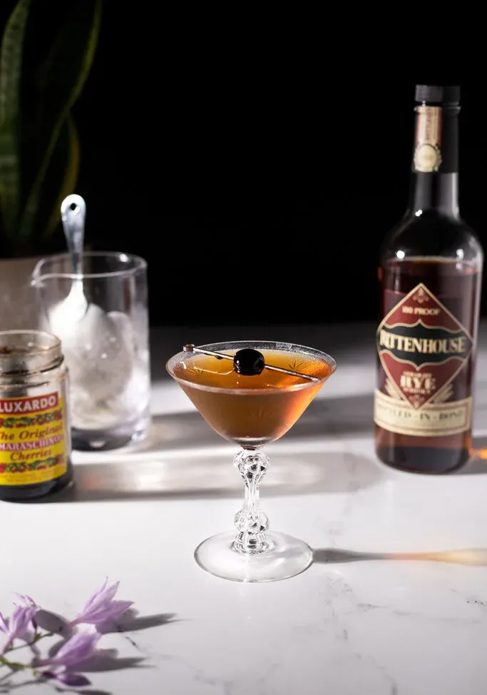 un cocktail marrone accanto a una bottiglia di whisky di segale e un bicchiere miscelatore.' loading='lazy' title=