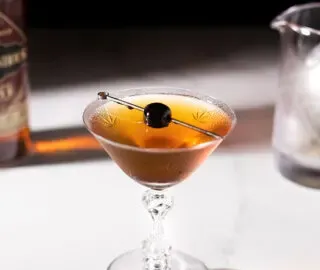 zabytkowy kieliszek koktajlowy wypełniony brązowym napojem i wiśnią na kilofie.' title=