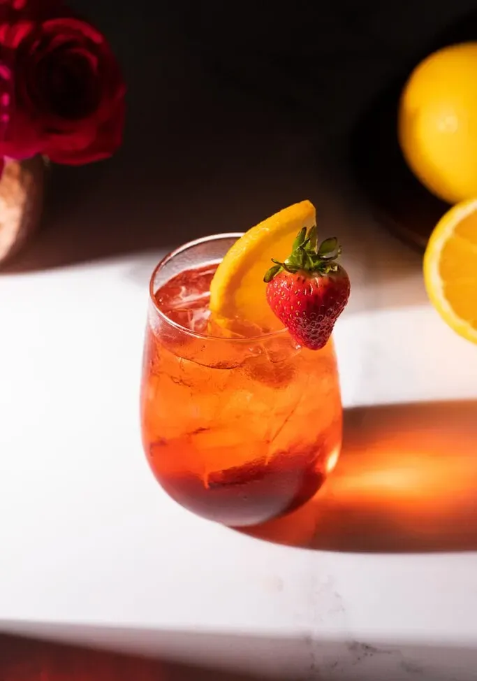 ein leuchtend rotes Getränk in einem stiellosen Weinglas mit einer Erdbeer- und Orangenscheibe.' loading='lazy' title=