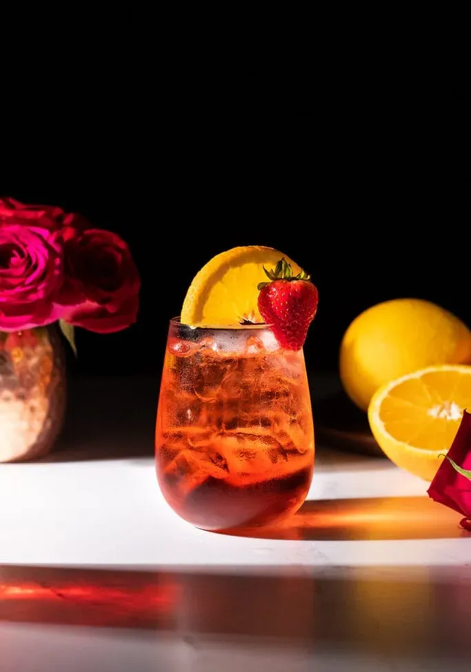 ein rotes Getränk in einem stiellosen Weinglas mit Orangenscheibe und Erdbeere.' title=