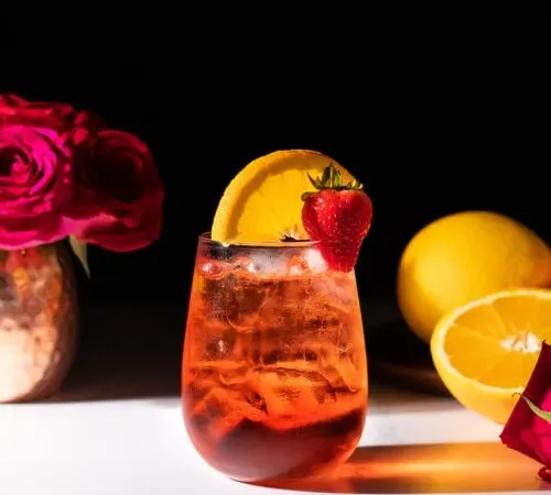 ein rotes Getränk in einem stiellosen Weinglas mit Orangenscheibe und Erdbeere.' title=