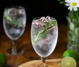 kielich wypełniony przezroczystym, musującym napojem z rozmarynem i różowymi ziarnami pieprzu.' title=