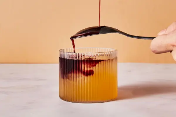 Une main tenant une cuillère à l'envers sur un verre à whisky, avec du vin rouge qui se verse lentement sur la cuillère et dans le verre à whisky