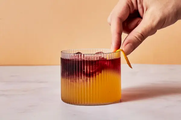 Une main qui passe un zeste de citron sur le bord d'un New York Sour dans un verre à whisky