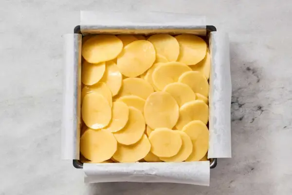 Uma assadeira quadrada forrada com papel manteiga e coberta com batatas fatiadas com gordura de pato