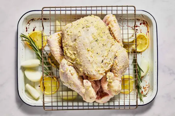 Un pollo entero untado con mantequilla con hierbas sobre una rejilla para cocinar, sobre una fuente para asar con limones, rodajas de cebolla y hierbas frescas.