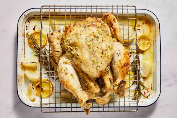 Un pollo dorado con hierbas enteras untado con mantequilla sobre una rejilla para cocinar, sobre una fuente para asar con limones, rodajas de cebolla y hierbas frescas.