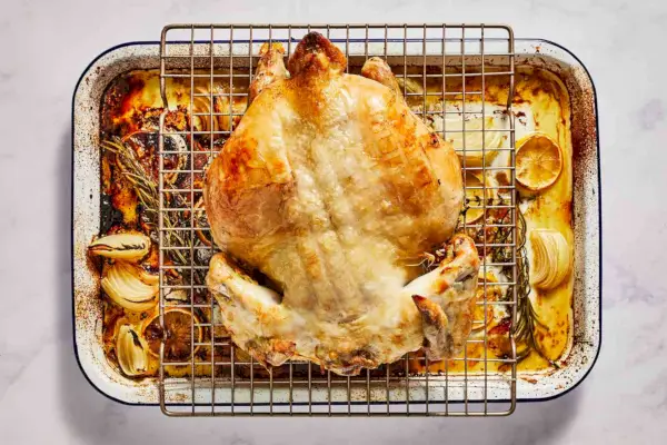 Un pollo dorado y untado con mantequilla de hierbas, con la pechuga hacia abajo, sobre una rejilla para cocinar, colocado sobre una fuente para horno con limones, cebollas y hierbas.