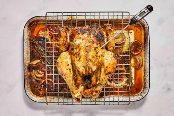 Un pollo asado untado con mantequilla de hierbas sobre una rejilla para cocinar, colocado sobre una fuente para hornear con limones, cebollas y hierbas, con un termómetro de cocina