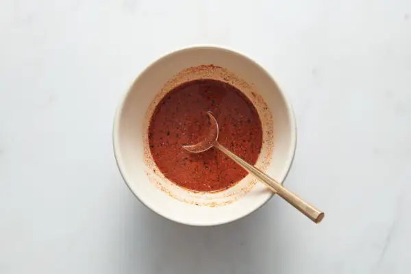Un tazón pequeño con salmuera de pepinillos, sal, pimentón, pimienta negra, pimienta de cayena, ajo en polvo y cebolla en polvo que se revuelve con una cuchara.