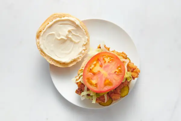Un sándwich de pollo frito con pepinillos, muslos de pollo frito, lechuga rallada, una rodaja de tomate y mayonesa en un panecillo.