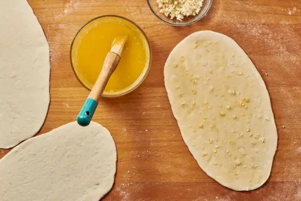 Una tabla de cortar enharinada con un trozo de masa aplanado cubierto con ajo picado y untado con mantequilla.